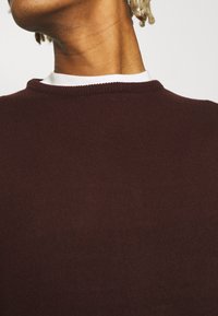 Burgundyröd stickad tröja med ribbad struktur och rund halsringning, lagd över en vit skjorta. Närbild av nacke och axel.