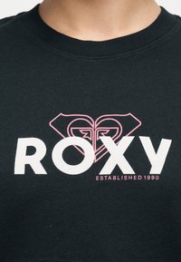 Μαύρο πουκάμισο με λευκή επιγραφή "ROXY" και ροζ λογότυπο σε σχήμα καρδιάς πάνω από την επιγραφή "Established 1990" τυπωμένα στο στήθος, που φοριέται από ένα άτομο.
