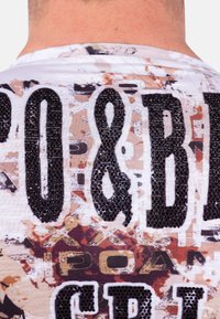Witte graphic t-shirt met grote zwarte letters bezaaid met rhinestones, op een kleurrijke gestructureerde achtergrond van abstracte ontwerpen en patronen.