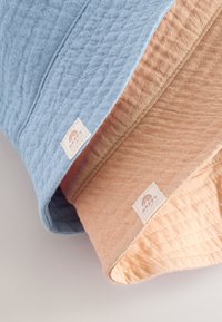 Divi teksturēti bļodas cepures gaiši zilā un bēšā krāsā ar nelielām auduma etiķetēm, kurās redzams varavīksnes logo, parādītas vienai uz otras uz gaišas virsmas.