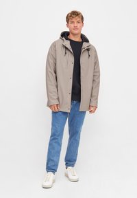 Light gray waterproof jacket with a hood, featuring black drawstrings, front snap closure, and a relaxed fit, worn over a black shirt.