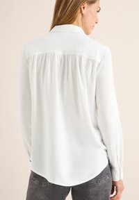 Blouse blanche à manches longues avec une texture douce, un col plié et un détail plissé dans le dos, associée à un jean en denim gris.