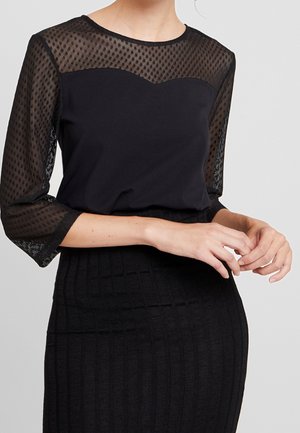 Kobieta ubrana w czarny top z przezroczystymi, kropkowanymi rękawami i karczkiem z siateczki, w połączeniu z czarną, prążkowaną spódnicą z wysokim stanem.