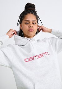Felpa grigia in materiale felpato con tasca frontale, caratterizzata da un logo "Carhartt" ricamato in bordeaux e cappuccio con cordoncino.