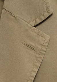 Primo piano di un tessuto beige che mette in evidenza una texture liscia, un colletto ben definito e una tasca con dettagli di cucitura visibili.