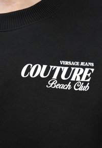 Μαύρο ύφασμα με λευκό κείμενο "Versace Jeans Couture Beach Club" τυπωμένο κοντά στον γιακά αριστερά.