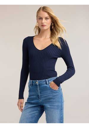 Kvinde med blondt hår iført en navy ribbet langærmet top og lyseblå jeans, poserer med den ene hånd i lommen mod en neutral baggrund.