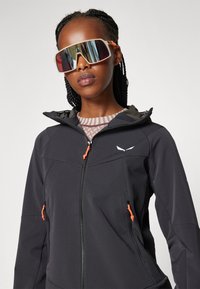 Čierna softshell bunda s oranžovými akcentmi, so zipsovými vreckami, kapucňou a bielym logom na hrudi. Model má na sebe reflexné slnečné okuliare.