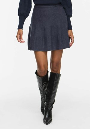 Femme portant une jupe trapèze gris foncé, un haut à manches longues noir et des bottes en cuir noires pointues jusqu'aux genoux, debout devant un fond blanc.