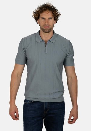 Hombre con el pelo rizado que lleva un polo de punto gris claro de manga corta con cuello de cremallera y vaqueros azul oscuro, de pie sobre un fondo liso.