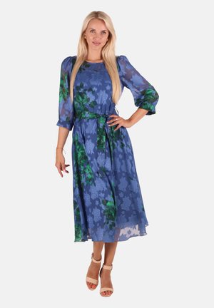 Blaues florales Midi-Kleid mit durchsichtigen Ärmeln, grünen Akzenten, Taillengürtel und weichem Material. Kombiniert mit beigen Sandalen mit offener Zehenpartie.