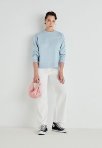 Lichtblauwe sweatshirt met geribbelde manchetten en zoom, witte tekstlogo, gecombineerd met witte wijde broek en zwarte hoge sneakers, met een roze tas.