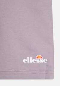 Lilla kortermet skjorte laget av mykt stoff, med et trykt hvitt "ellesse"-logo og en fargerik aksent over teksten.