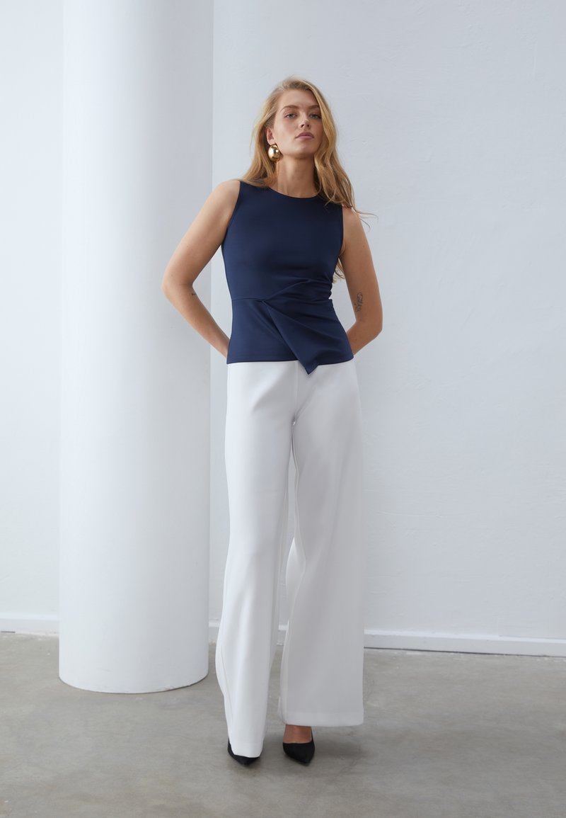 Granatowy bezrękawnik z skręcanym przodem, zestawiony z białymi spodniami o prostych nogawkach i wysokim stanem oraz czarnymi czółenkami.