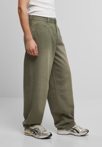Pantalones de algodón verde oliva claro con un corte relajado, que cuentan con cinco bolsillos y una textura suave. Combinados con zapatillas deportivas plateadas y beiges.
