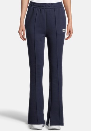 Donkerblauwe fleecebroek met elastische tailleband, verticale naden, klein logo-patchje op de linker dij, gedragen met witte sneakers.