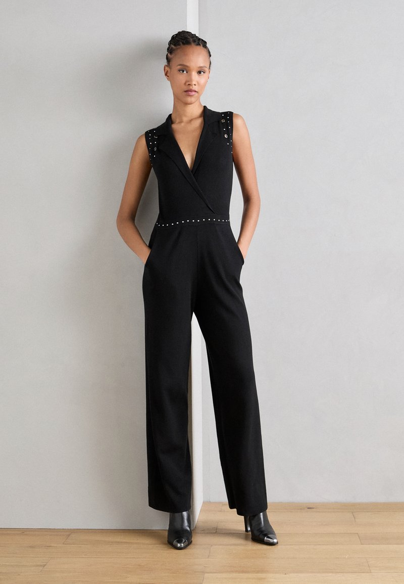 Combinaison sans manches noire avec un décolleté en V profond, des épaules ornées et un détail à pression à la taille ; coupe ajustée avec des jambes larges.