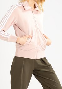 Rosa zip-up jacka med vita ränder på ärmarna, framtida fickor och en mjuk, slät textur. Matchad med lösa olivgröna byxor.