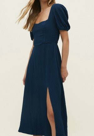 Vrouw draagt een midijurk van marineblauw met een gestructureerde stof, pofmouwen, een vierkante halslijn en een hoge split aan de linkerkant.