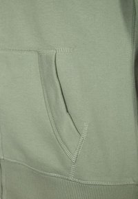 Světlé zelené mikina s přední kapsou a žebrovaným lemem. Měkká látka s viditelným švem. Hladká textura.