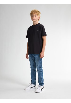 Schwarzes Kurzarm-T-Shirt mit einem kreisförmigen Logo auf der Brust, kombiniert mit blauen Denim-Jeans und weißen Sneakern mit schwarzen Akzenten.