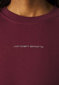 Camiseta de cuello redondo en color burdeos, fabricada con material acanalado, que presenta un pequeño logo plateado de "jordan sport" en el frente.