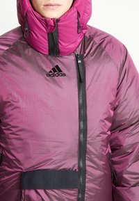 Magenta puffjacka med hög krage, svart dragkedja och justerbara dragsnören. Har en Adidas-logotyp och texturerad tyg.