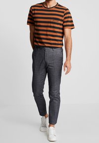 Hombre vestido con una camisa de manga corta a rayas naranja y negra, pantalones grises ajustados y zapatillas blancas, de pie contra un fondo liso.