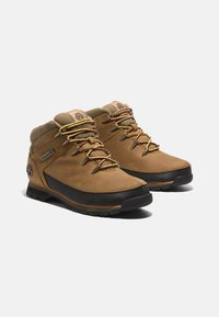 Braune Wanderstiefel aus Leder mit schwarzen Gummisohlen, goldenen Schnürsenkeln, gepolsterter Blende und seitlichem Logo; für den Einsatz im Freien konzipiert mit robuster Bauweise.