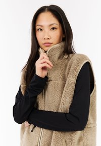 Gilet in pile beige con colletto alto, chiusura con zip e design senza maniche. Sotto, una maglietta nera a maniche lunghe; le trame sono messe in risalto dal materiale morbido.