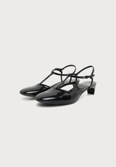 Paire de chaussures slingback en cuir verni noir à petit talon avec bride en T et boucle latérale sur fond blanc.