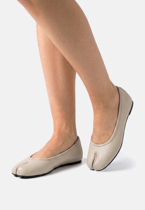 Nogi w beżowych płaskich butach z rozdzielonym palcem i czarnymi podeszwami stojące na białej powierzchni.