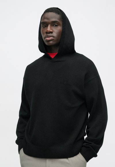 Bărbat îmbrăcat într-un pulover cu glugă negru, cu mâinile în buzunare, stând pe un fond simplu deschis la culoare, privind înainte cu o expresie neutră.