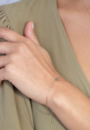 Gold chain bracelet with a small heart charm, worn on a hand against a light green fabric background. The chain features delicate links.