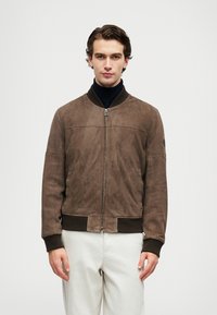 Young man wearing a brown suede bomber jacket with ribbed cuffs and collar, black turtleneck, and light beige pants, standing against white background.