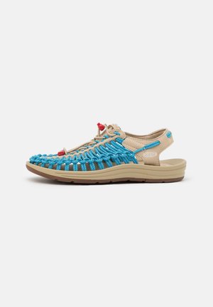 Beige sandal med vævede klare blå snore, justerbare elastiske snørebånd, polstret sål og åben hæl til udendørs eller afslappet brug.
