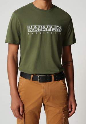 Man wearing olive green short-sleeve Napapijri graphic T-shirt, beige cargobroek en zwarte riem, staand tegen een effen achtergrond.