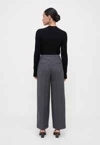 Vrouw met donker haar in een bun, gekleed in een zwarte lange mouwen top, grijs gestreepte wijde broek en zwarte sandalen met hak, staande met de rug naar de kijker.