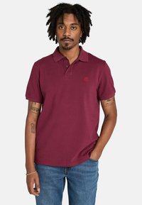 Un joven con cabello rizado está de pie, vistiendo una camiseta polo burdeos y jeans, con los brazos relajados a los lados y una expresión neutral.
