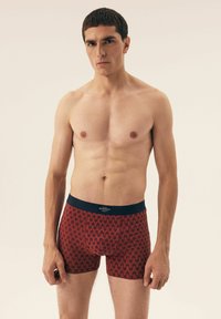 Red patterned boxer shorts with a navy waistband. The fabric appears soft with a wavy design, fitted to the model's body.