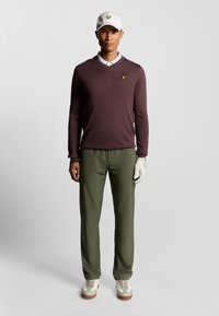 Outfit da golf composto da un maglione a maglia bordeaux, pantaloni verde oliva, cappellino bianco con logo e scarpe chiare. Guanti sulla mano sinistra.