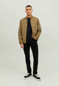 Jovem vestido com uma jaqueta bomber bege, camisa preta, calças pretas e ténis pretos com solas e atacadores brancos, de pé com uma mão no bolso.