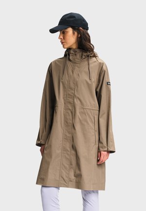 Femme portant un imperméable à capuche beige et une casquette de baseball noire, debout de profil avec les mains détendues le long du corps sur un fond uni.