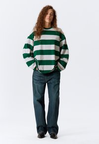 Zeleno-sivý pásikavý oversized sveter s širokými rukávmi, skombinovaný s voľnými tmavými džínsami a čiernou obuvou. Jednoduchý dizajn.