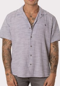 Camisa de manga corta con botones hecha de una tela ligera de color púrpura texturizado, que presenta un patrón sutil y un pequeño logo bordado en el lado izquierdo del pecho.
