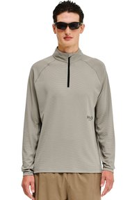 Hellgraues, langärmeliges Oberteil mit Reißverschlusskragen, gefertigt aus strukturiertem Stoff, mit schwarzem Reißverschluss und bedrucktem Logo auf der linken unteren Seite. Getragen mit Sonnenbrille.