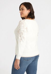 Maglione bianco con ricami floreali in rosa oro sulle maniche, polsini e orlo a coste, abbinato a jeans blu. Morbida texture in maglia in tutto il capo.