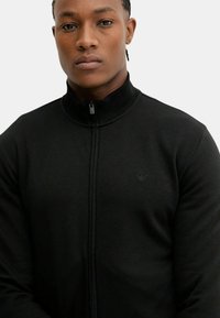 Svart zip-up hoodie med hög krage, tillverkad av släta tyger. Har en liten logotyp på bröstet och ribbade detaljer vid fållen.