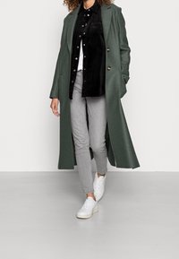 Cappotto lungo verde in lana con revers a punta, abbinato a una camicia nera in corduroy, jeans grigi e sneakers bianche.