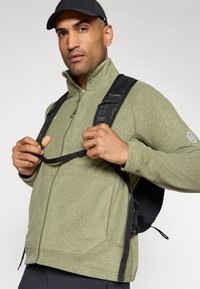Giacca in fleece verde oliva con colletto zip e dettagli di cucitura, indossata con uno zaino nero dotato di spallacci imbottiti.
